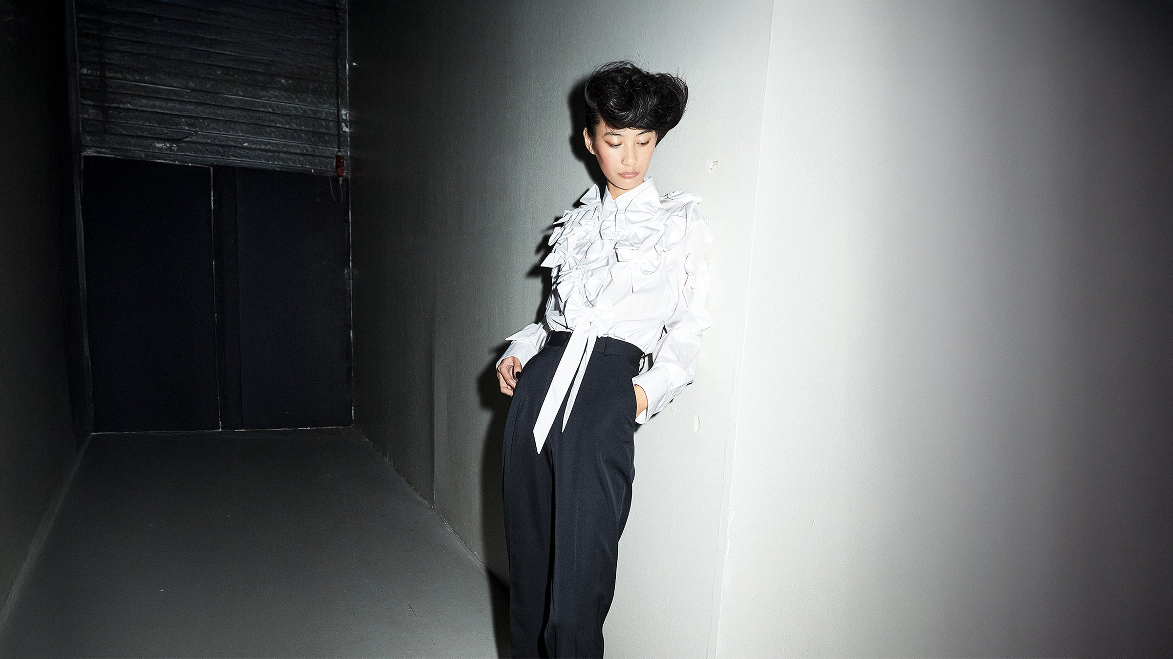 Woman standing against grey wall wearing a white shirt and black pants.