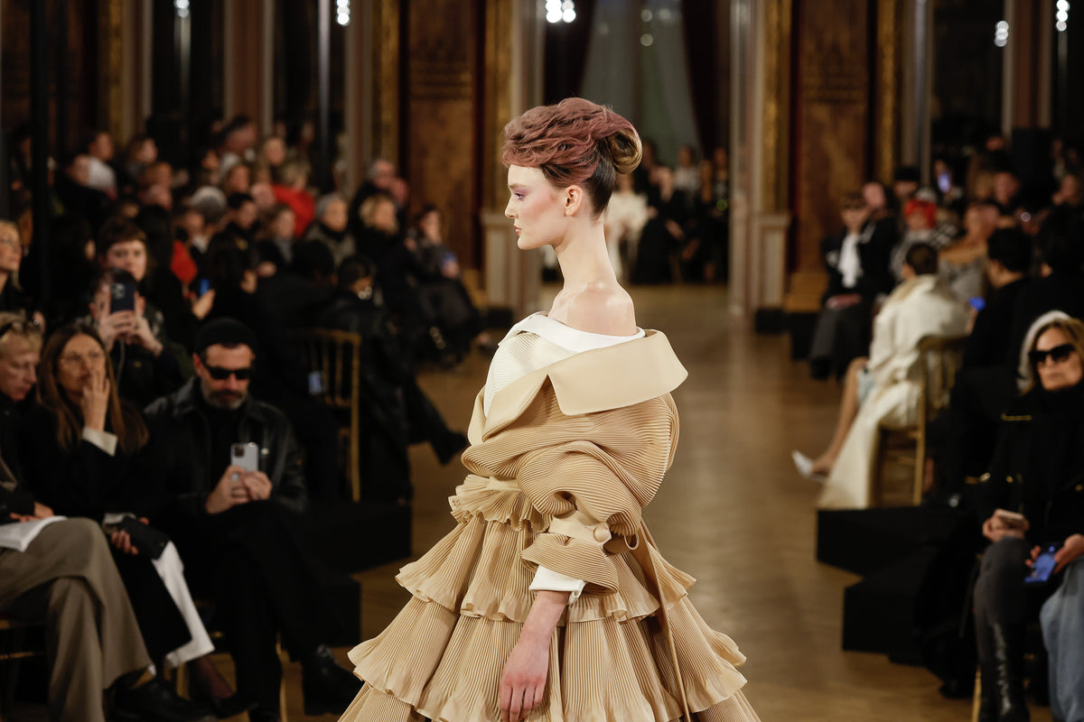 Model wearing a beige dress on a runway with an audience in a formal setting