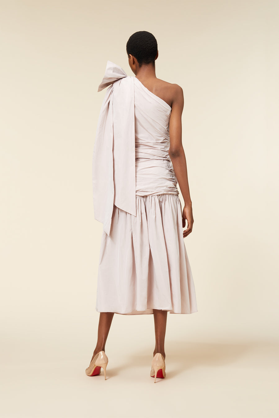 Elegant asymmetrical white dress with large shoulder bow, ruched bodice, and flared skirt, worn with beige high heels.