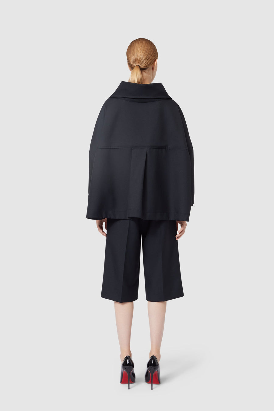 Back view of a woman in a stylish black oversized jacket and culottes, with patent high-heeled shoes on a light gray background.