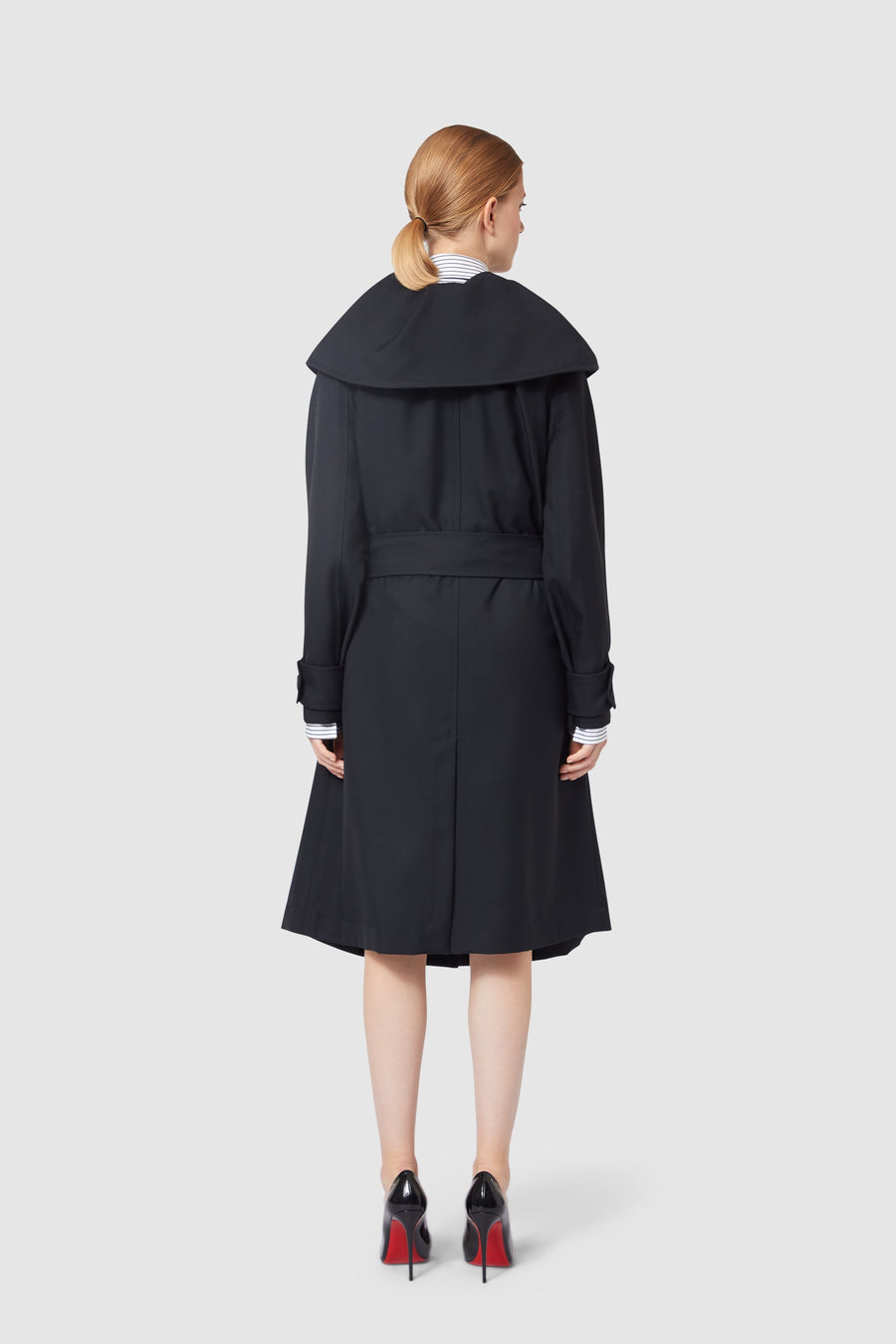 Elegant black wool coat with oversized collar, belted at the waist, over a striped shirt, paired with chic black stilettos.