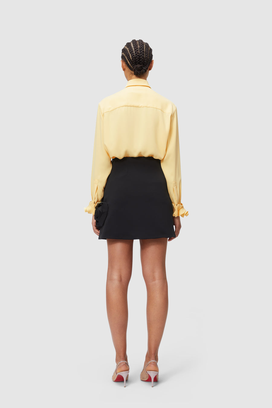 Yellow silk blouse with bow-tie cuffs paired with a black mini skirt and elegant red-soled stilettos, seen from the back.