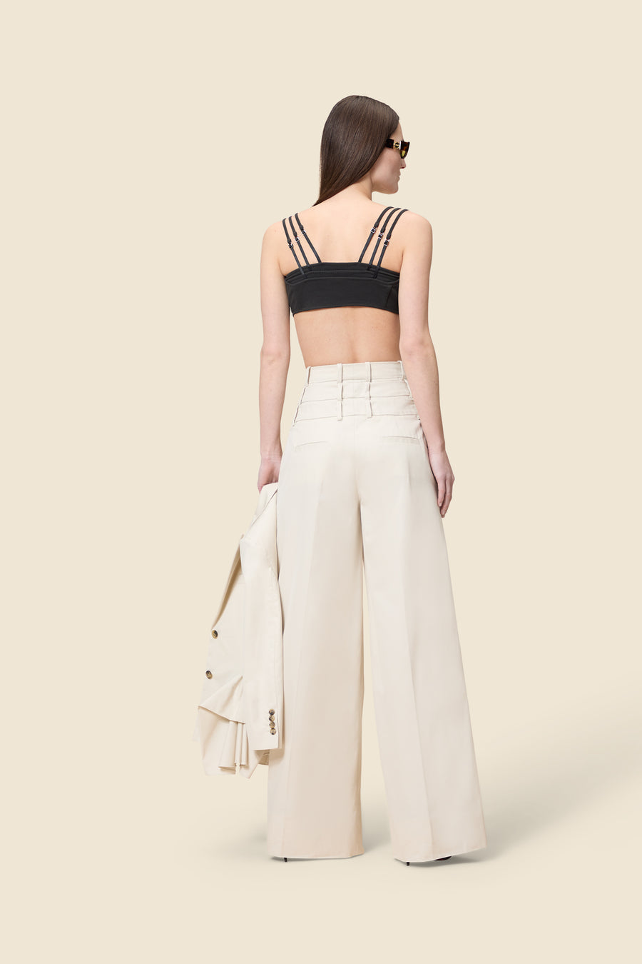 Model carrying a beige blazer with ruffles at the bottom, styled with black bralette top and high waist multi-layer beige trousers, standing on a beige background.