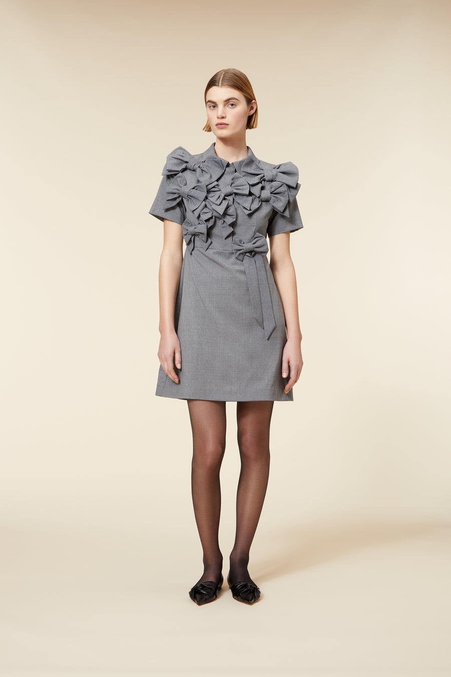 Gray dress with oversized bow accents, featuring short sleeves and knee-length skirt. Model wearing sheer black tights and flats.