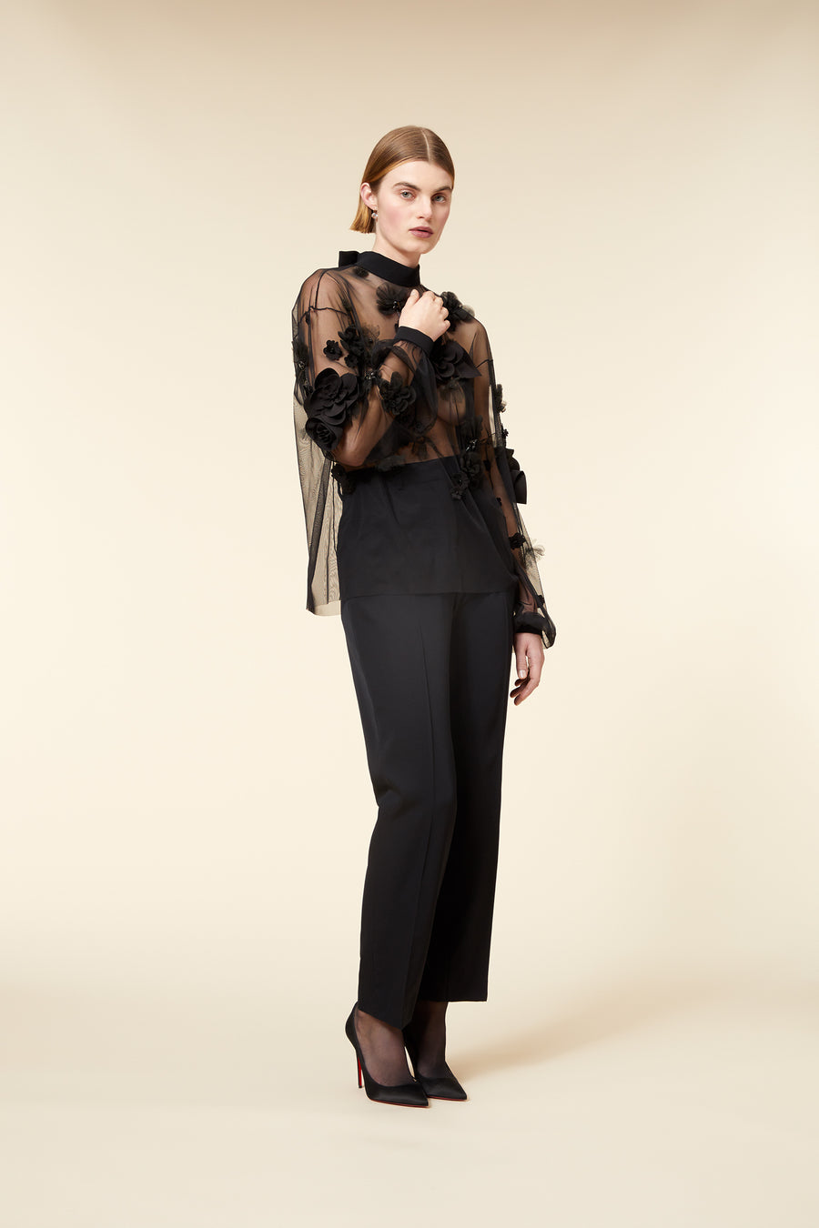 Model wearing a black sheer blouse with floral appliqué and matching trousers, posing against a neutral background.