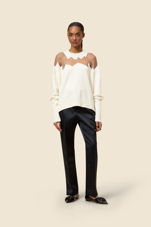 Model wearing a cream sweater with sheer zigzag design and sleek black straight-leg trousers on a plain background.