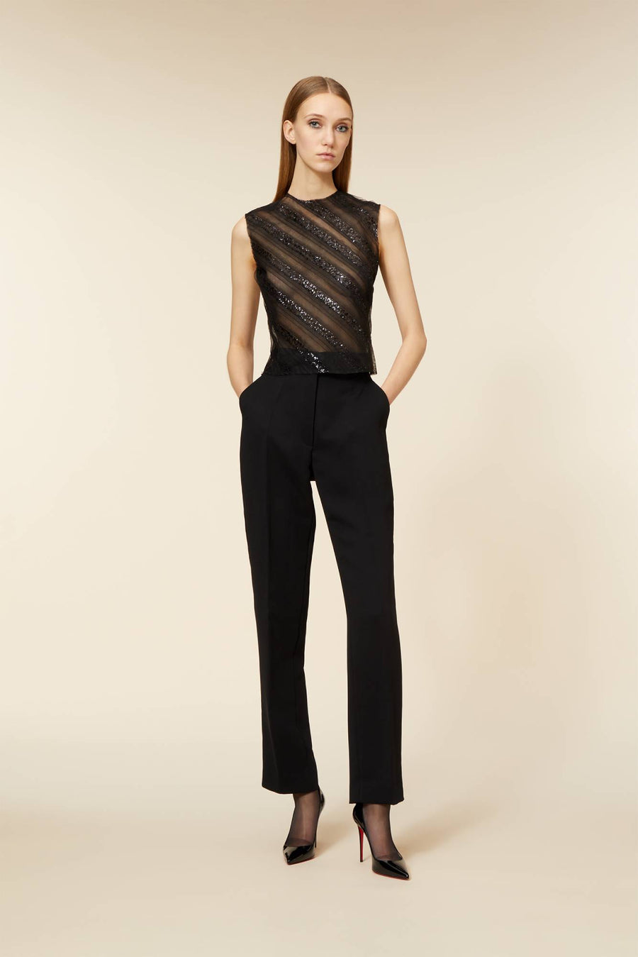 Elegant model wearing black sheer sleeveless top with sparkling stripes and black tailored trousers against plain background.