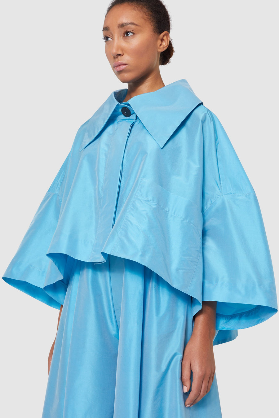 Woman wearing a vibrant blue oversized collared cape, featuring wide sleeves and a glossy finish.